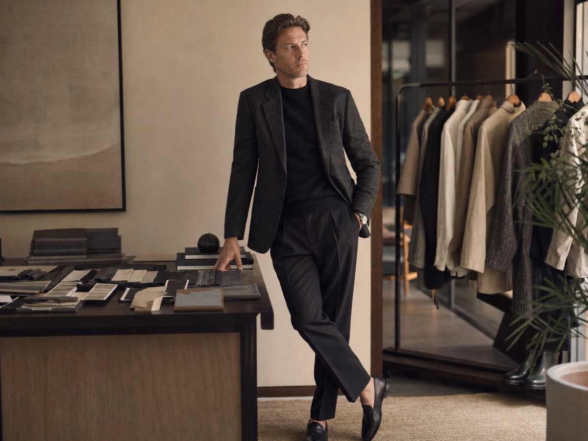Male executive in restrained dark tailoring beside a wardrobe rail and fabric swatches in a warm studio